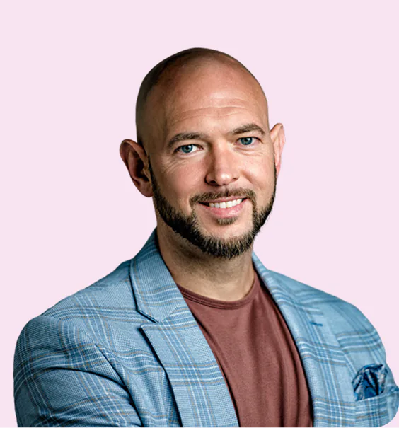 Smiling bald man with blue eyes and a short beard wearing a blue plaid blazer over a brown shirt, against a light purple background.